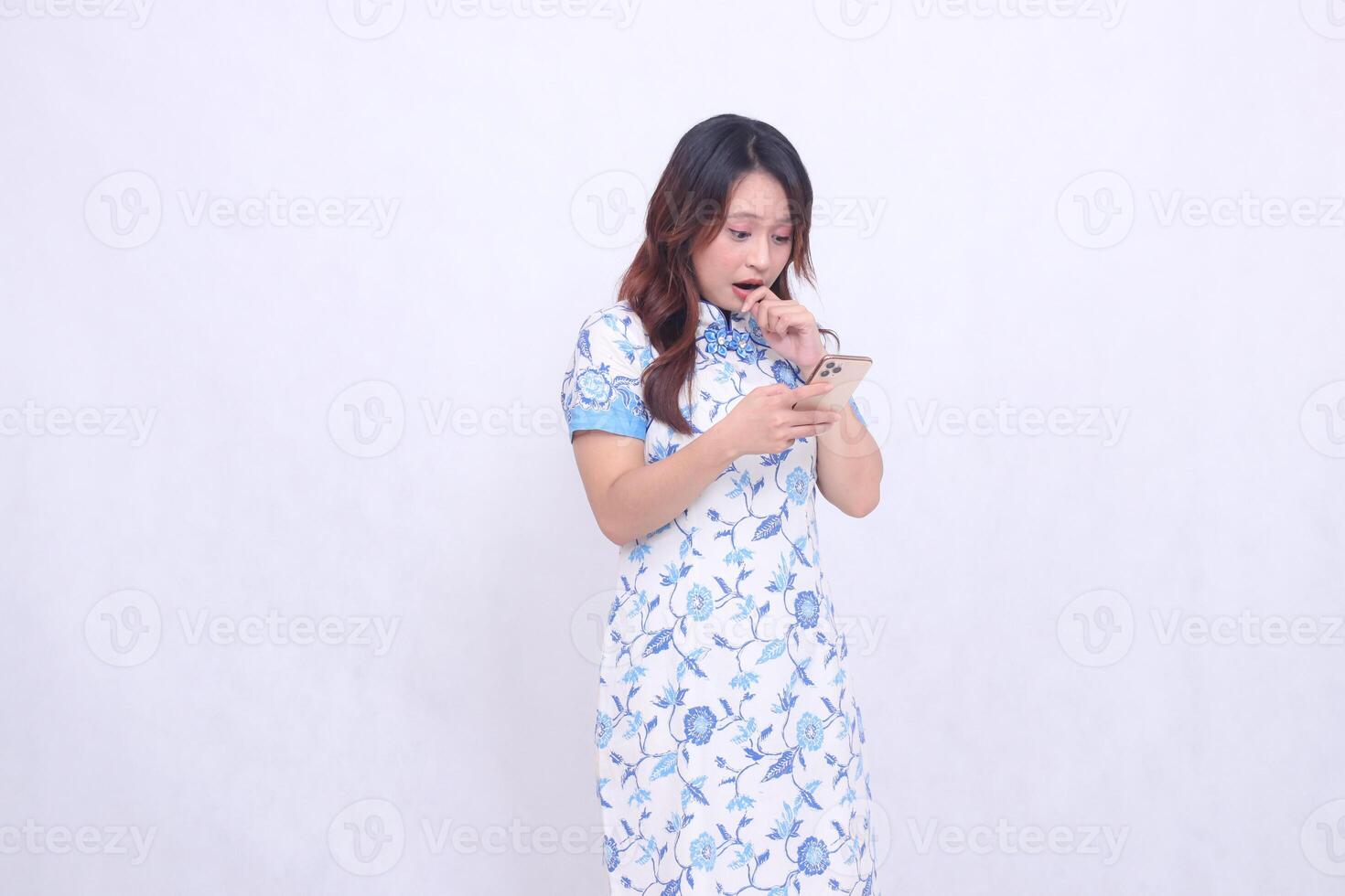 Young Asian Javanese Chinese woman wearing a blue Chinese dress is shocked to the left while focusing on typing on her cellphone isolated on a white background photo