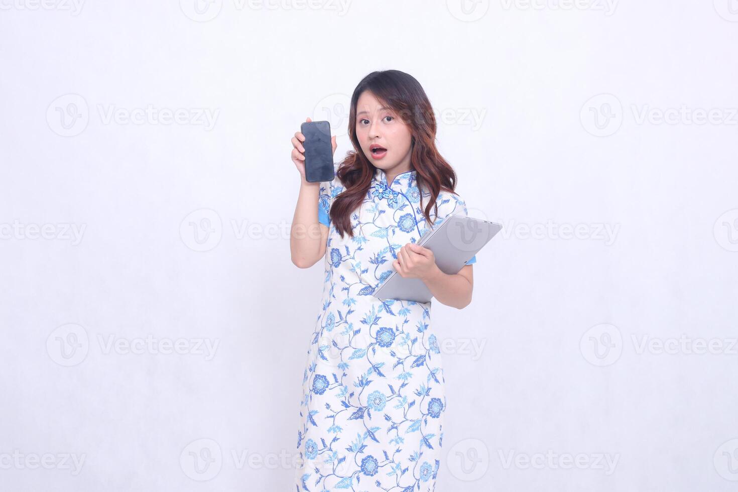 Beautiful Asian woman wearing blue Chinese dress holding tablet and looking at cellphone screen isolated on white background photo