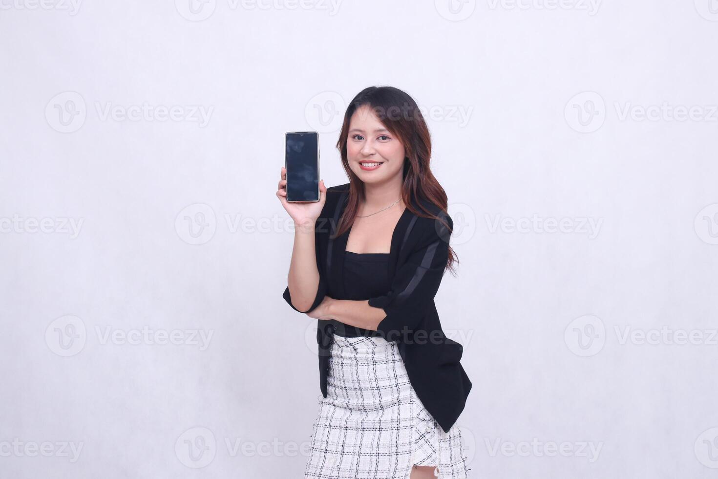 beautiful Indonesian office woman in her 20s smiling expression holding up cell phone gadget screen for promo while holding arms crossed isolated white background photo