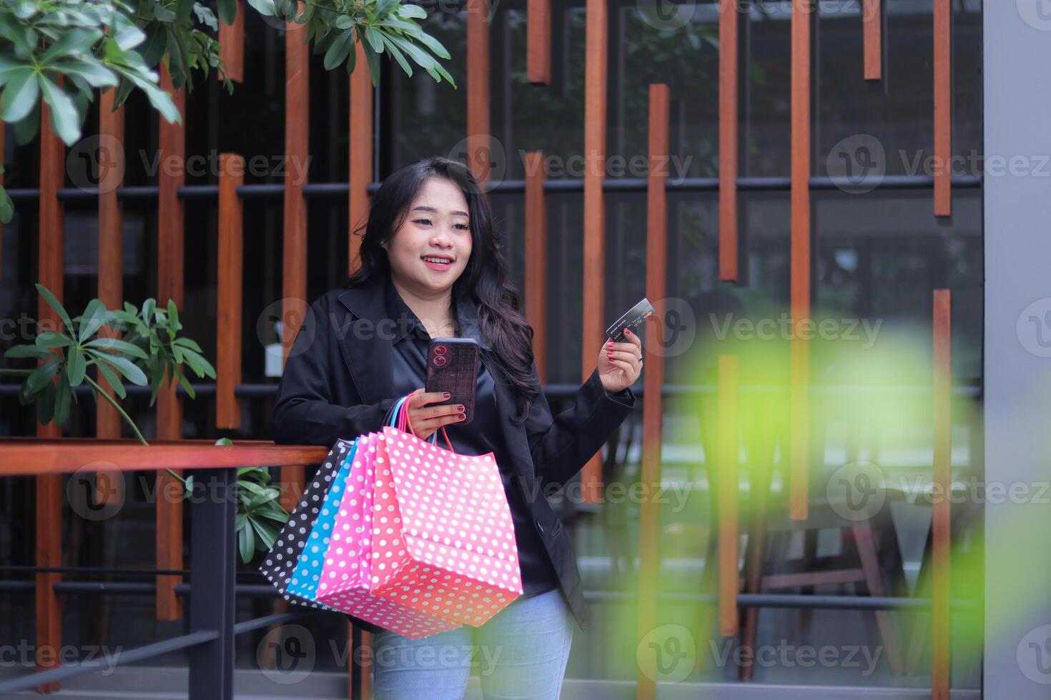 young Indonesian office woman with a candid smile shopping online to get a discount, standing against the fence carrying a paperbag and laptop computer, to promote online content, lifestyle, catalogs photo