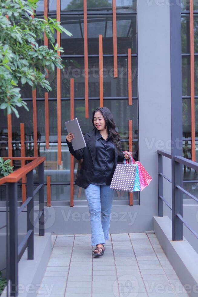 Cheerful young Indonesian female entrepreneur gets special prices for online orders, walks candidly, brings paperbags and laptop computers, to promote online content, lifestyle, catalogs photo