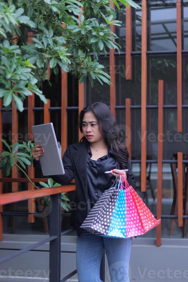 young asian businesswoman seriously checking reject orders, standing against fence carrying paperbag and computer laptop, used for online content promotion, lifestyle photo