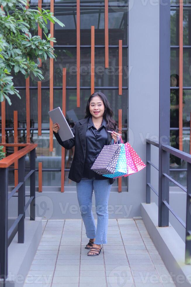 young Indonesian office girl is happy to get a discount price for an online order, walking candidly carrying a paper bag and laptop computer, to promote online content, lifestyle, catalogs photo