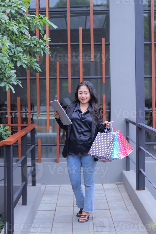 young Indonesian office girl is happy to get a discount price for an online order, looking at the camera, carrying a paper bag and laptop computer, to promote online content, lifestyle, catalogs photo