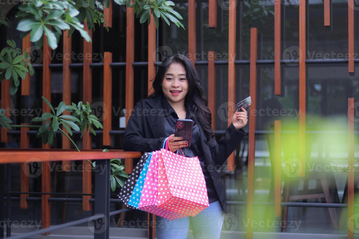 young Indonesian office girl smiling at the camera shopping online to get a discount, standing against the fence carrying a paperbag and laptop computer, to promote online content, lifestyle, catalogs photo