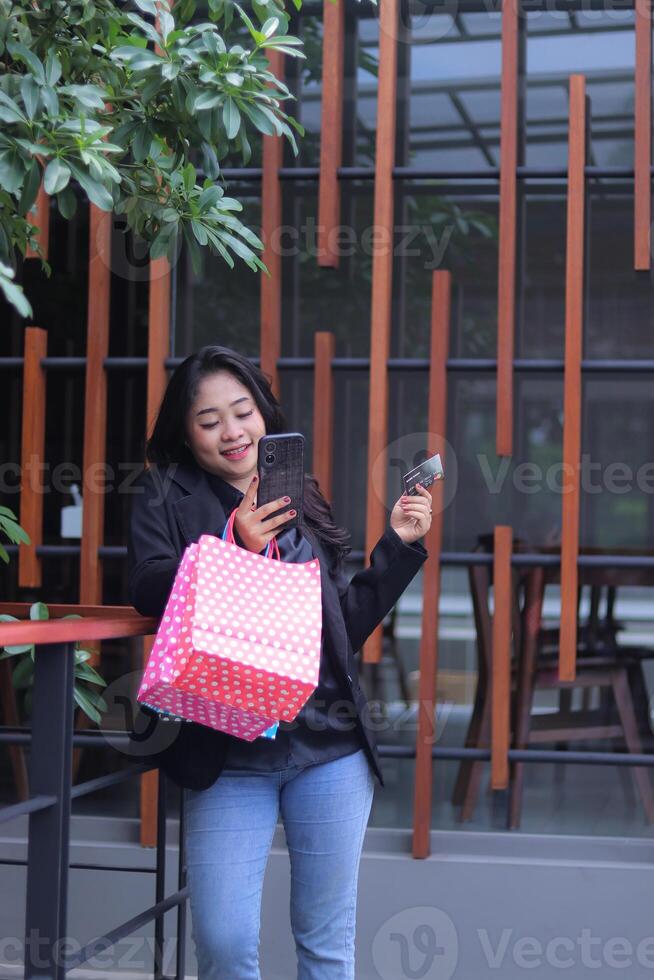 happy young Indonesian office woman checking online orders, standing against the fence carrying paperbags, credit cards and cell phones for promotion of online content, lifestyle, catalogs photo