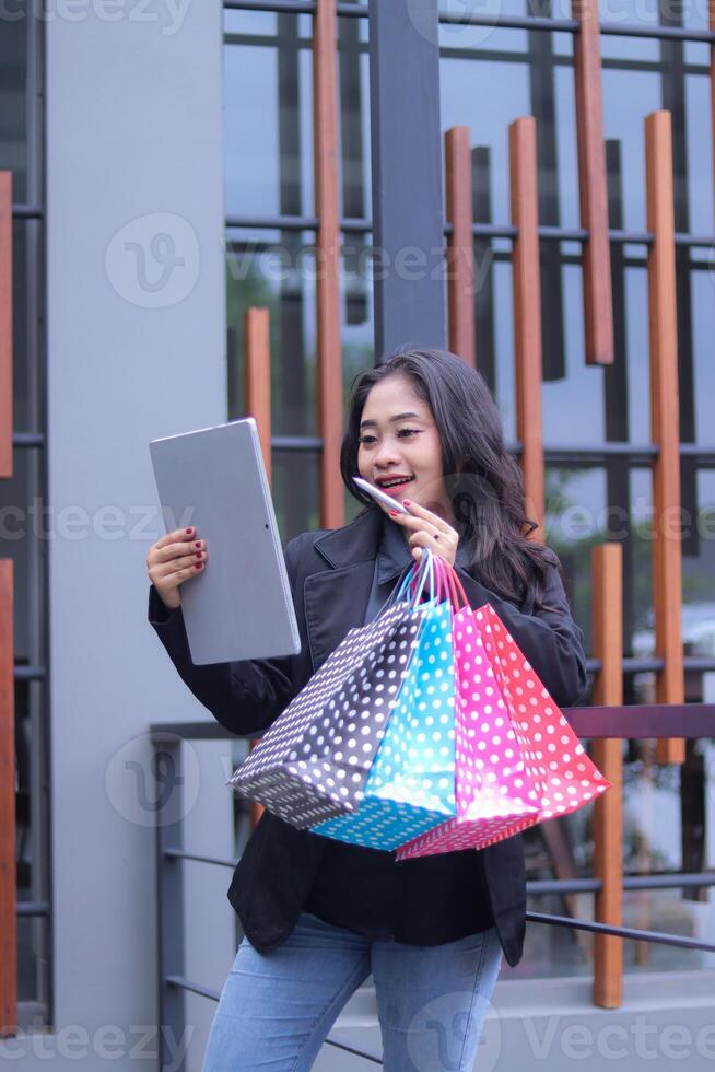 Young Indonesian businesswoman candidly happy to get discount on online order, standing against iron fence carrying paperbag and laptop computer, for promotion of online content, lifestyle, catalog photo