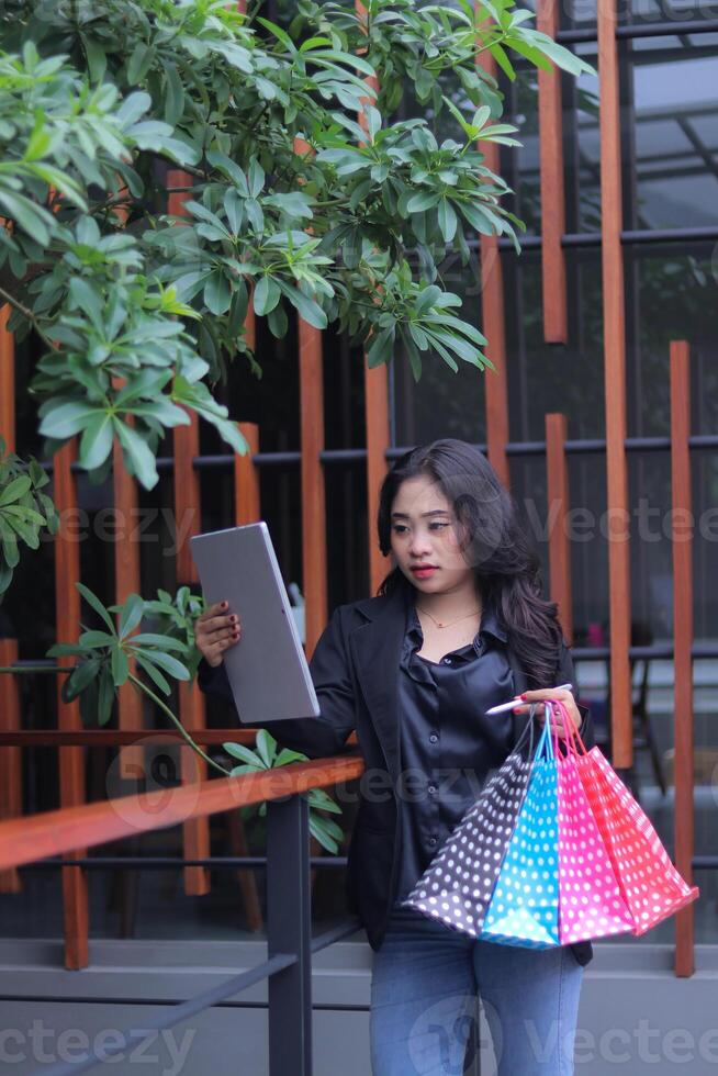 young asian businesswoman focused on checking orders, standing against fence carrying paperbag and computer laptop, used for online content promotion, lifestyle photo