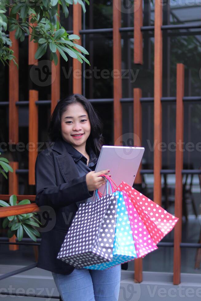 Cheerful young Indonesian office woman checking work, standing against the fence carrying a paperbag and laptop computer, for promotion of online content, lifestyle, catalog photo