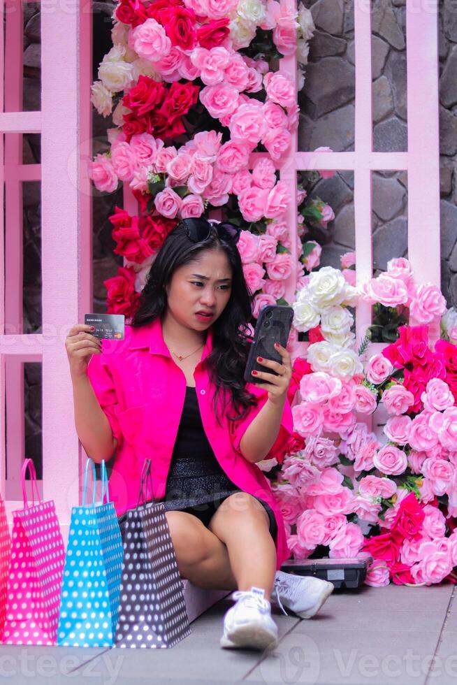 Beautiful Asian woman in pink shirt annoyed about checking out but connection error, sitting behind paperbag and credit card at outdoor cafe for promotion of fashion, lifestyle, catalog photo