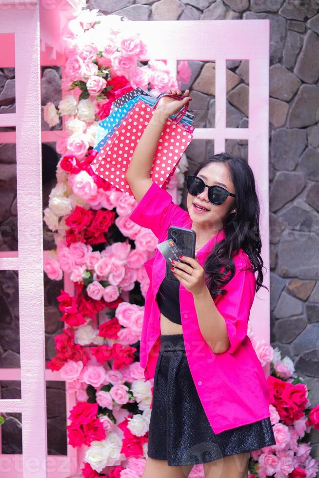 Beautiful Asian woman in pink shirt happy checking out on cellphone to get discount, standing carrying paperbag and credit card at outdoor cafe for promotion of fashion, lifestyle, catalog photo