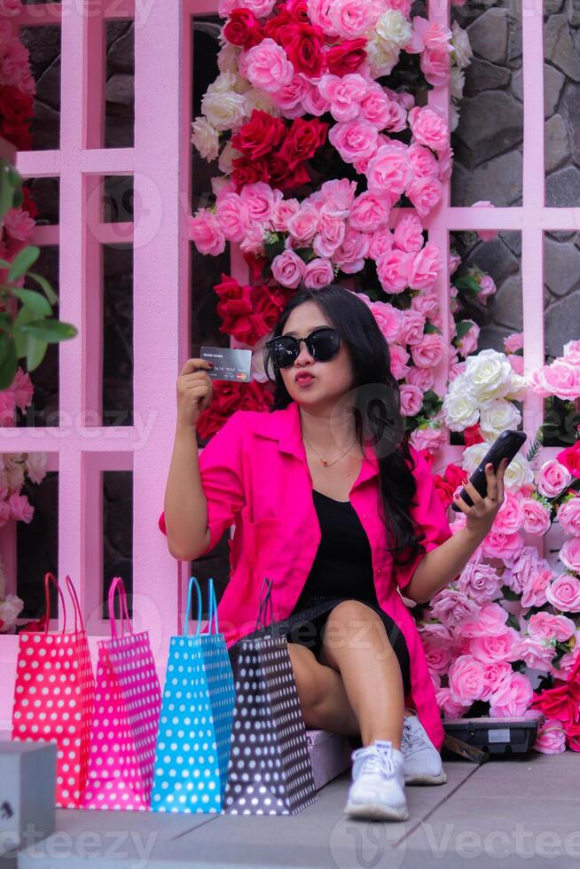 beautiful asian girl in pink shirt kissing wearing glasses happy hand holding debit card showing her shopping, sitting between paperbags in outdoor cafe for promotion of fashion, lifestyle, catalog photo