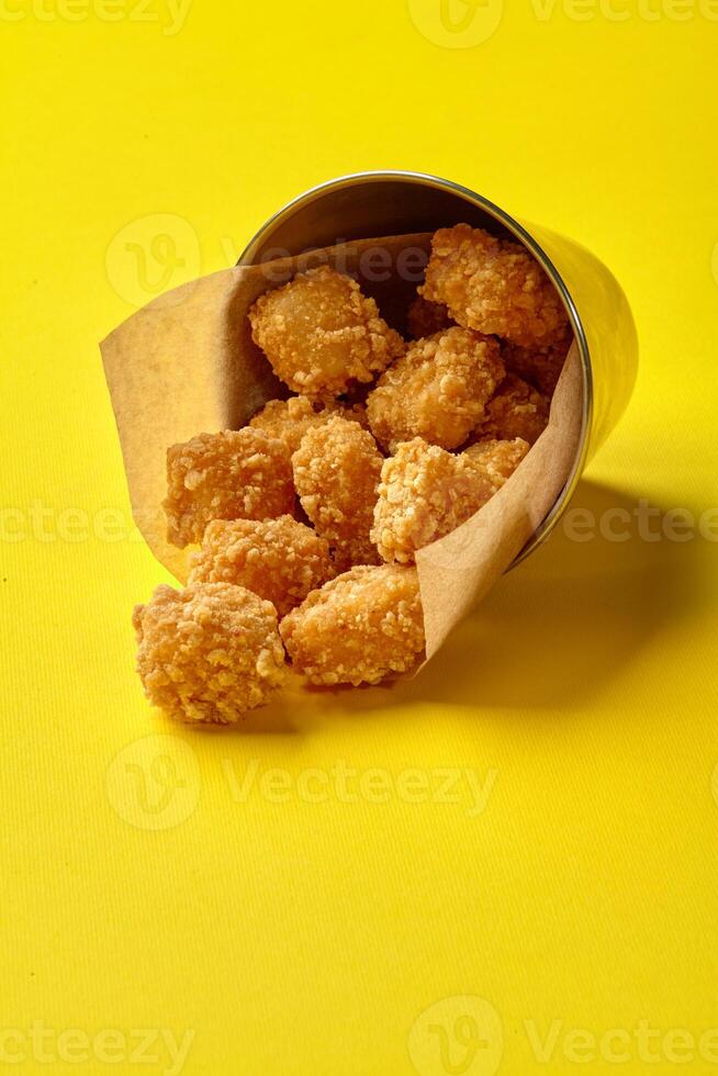 Crispy fried chicken nuggets spilling from metal bucket on yellow photo