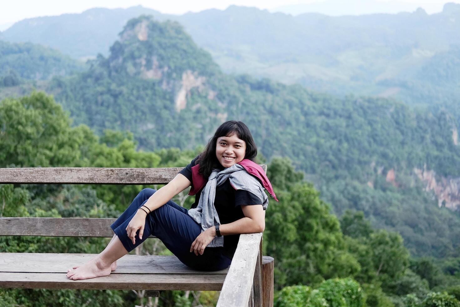 Asian woman sitting and relaxing on wooden bench on terrace for freedom journey with valley mountain and foggy in natural and travel destination in tropical rain forest photo