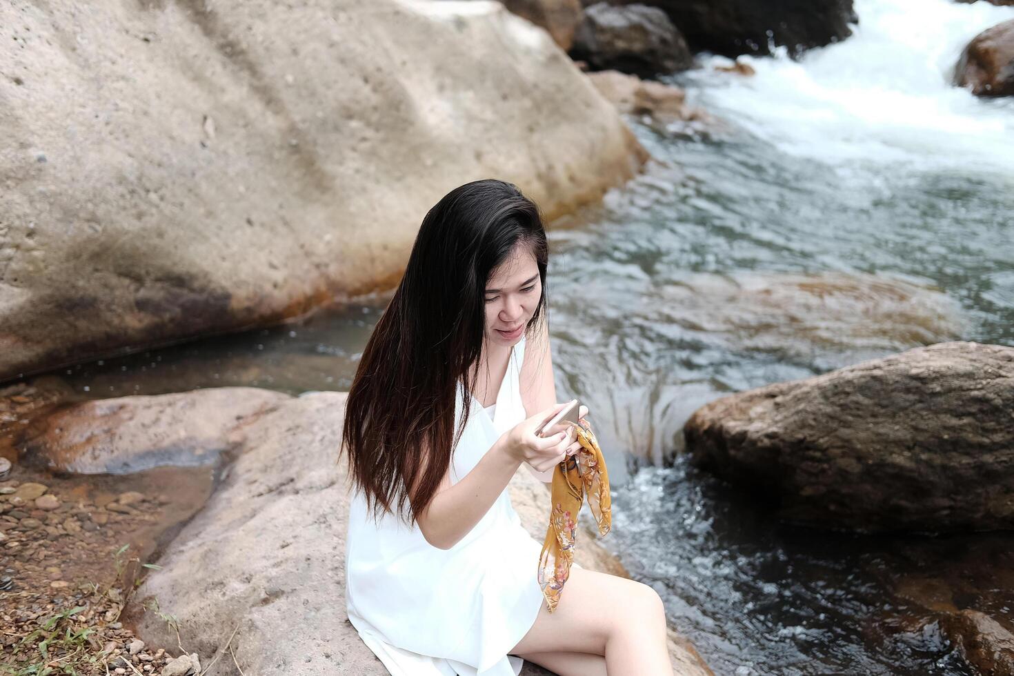 Smiling Beautiful asian female in white dress is sitting on the rock and relaxation for online shopping with samrtphone. Lifestyle of Technology for outdoor and stream in the tropical forest. photo
