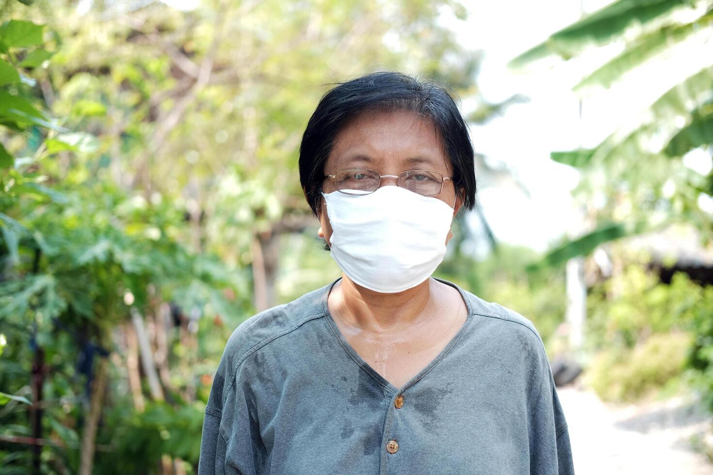 Asian old lady gardener is sweating and wearing glasses with white cloth mask for prevent the Covid-19 or Corona virus in Thailand and protect for Air Pollution Value Pm 2.5. Health elderly Concept. photo
