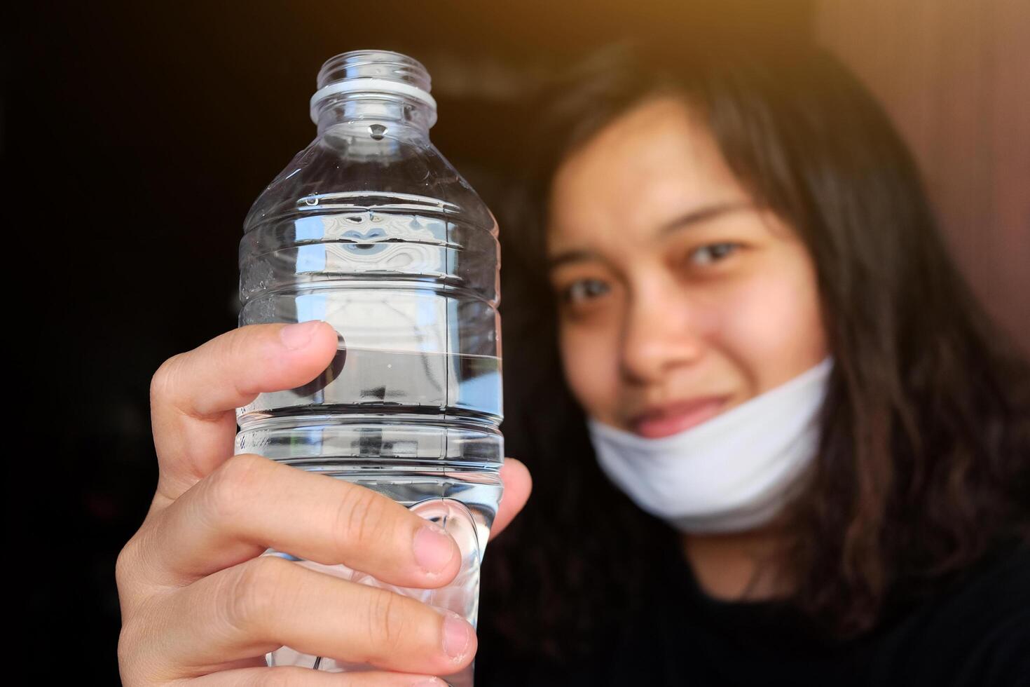 Asian Thai woman wearing a white cloth mask for prevent the Covid-19 or Corona virus and Air Pollution Value Pm 2.5 in Thailand and hand holding a bottled mineral water. Focus on Clear plastic Bottle. photo