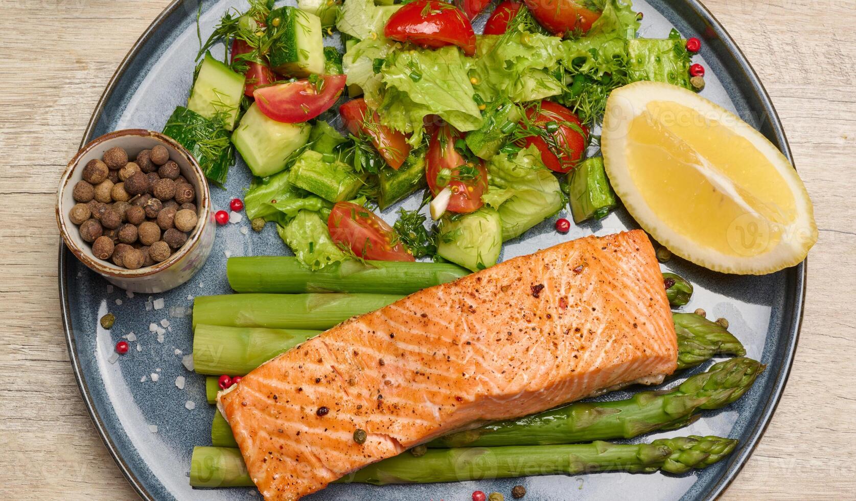 Healthy lunch with grilled salmon with asparagus and fresh tomato and cucumber salad, a row of lemon slice. Food on a blue round plate photo