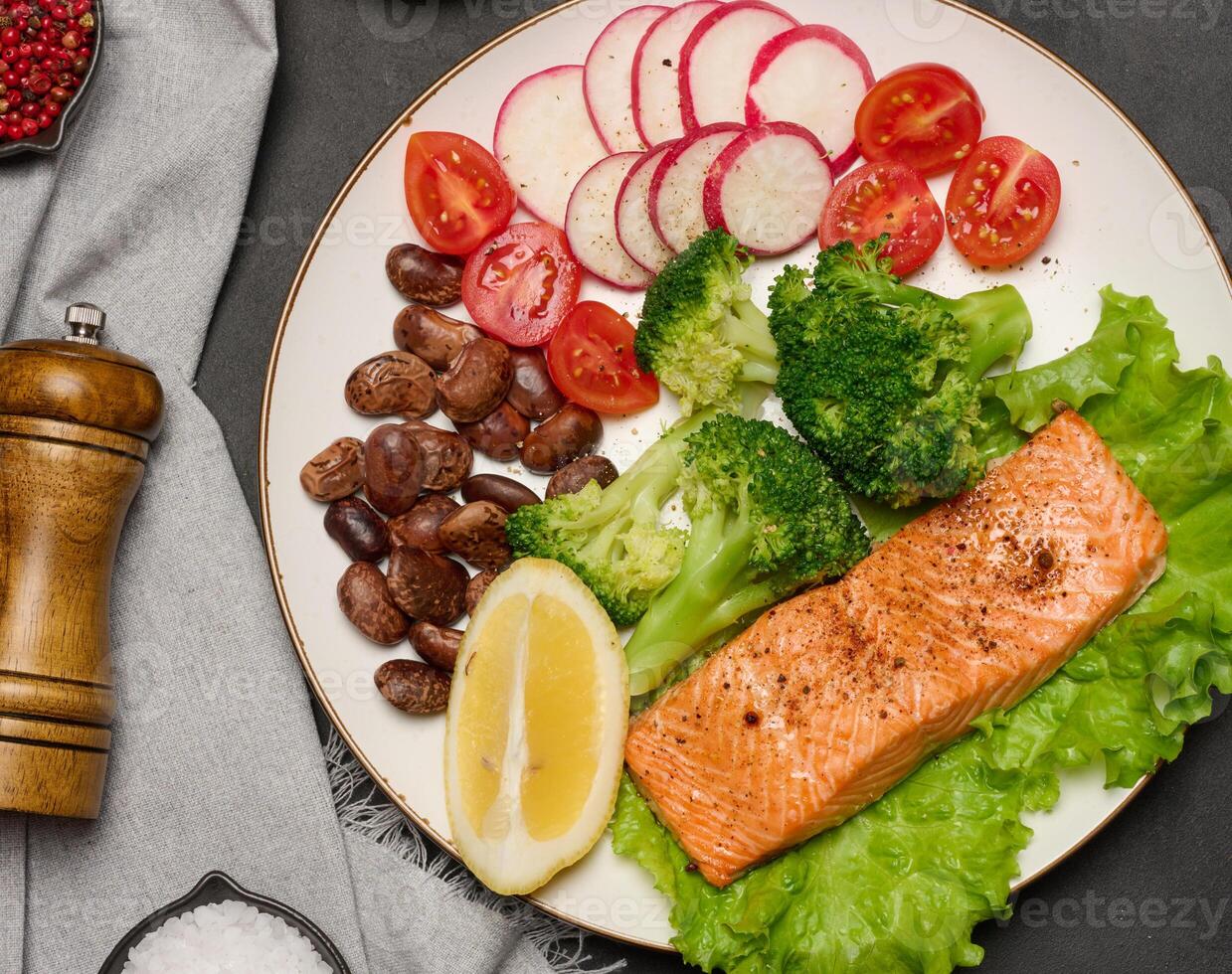 Healthy lunch with grilled salmon on green lettuce, next to vegetables, tomatoes, radishes, broccoli and a portion of beans. photo