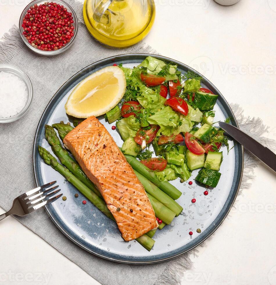 Healthy lunch with grilled trout with asparagus and fresh tomato and cucumber salad, a lemon slice. Food on a blue round plate photo