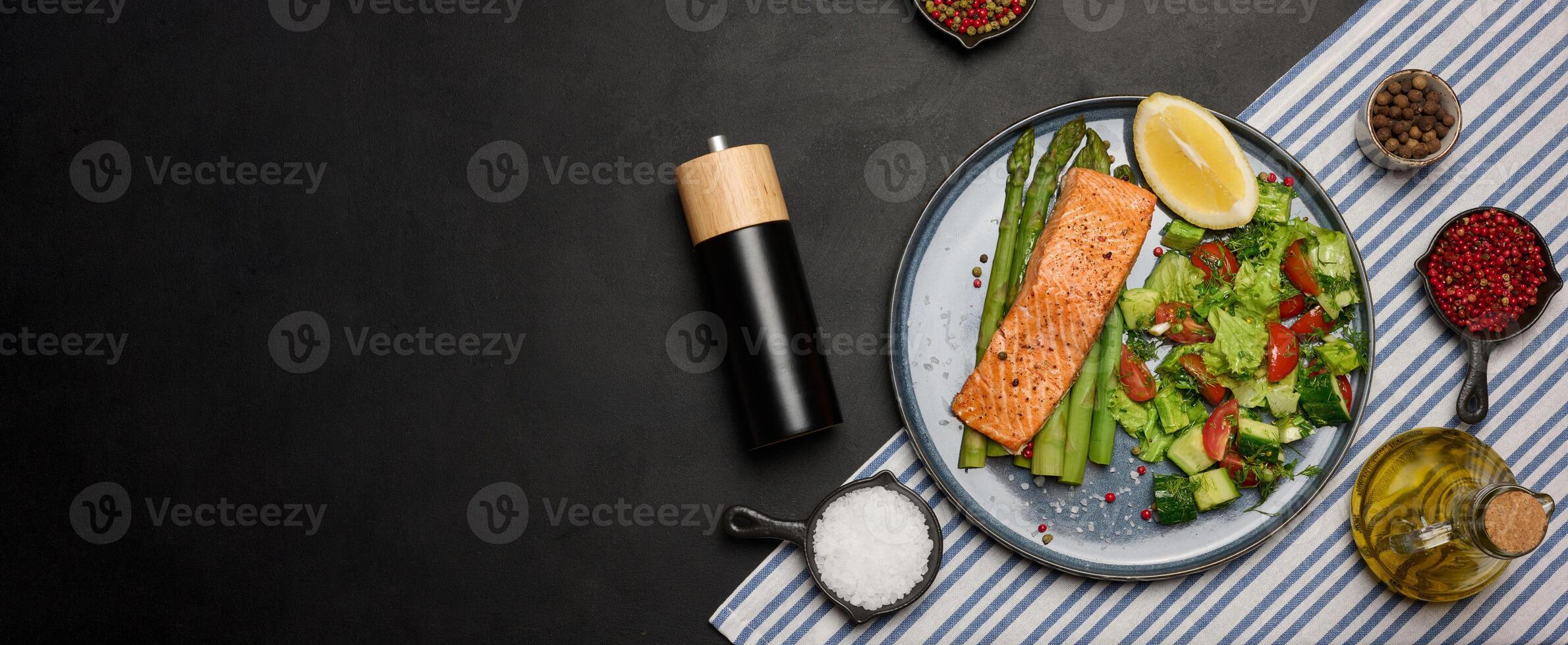 Healthy lunch with grilled trout with asparagus and fresh tomato and cucumber salad, a lemon slice. Food on a blue round plate, top view photo