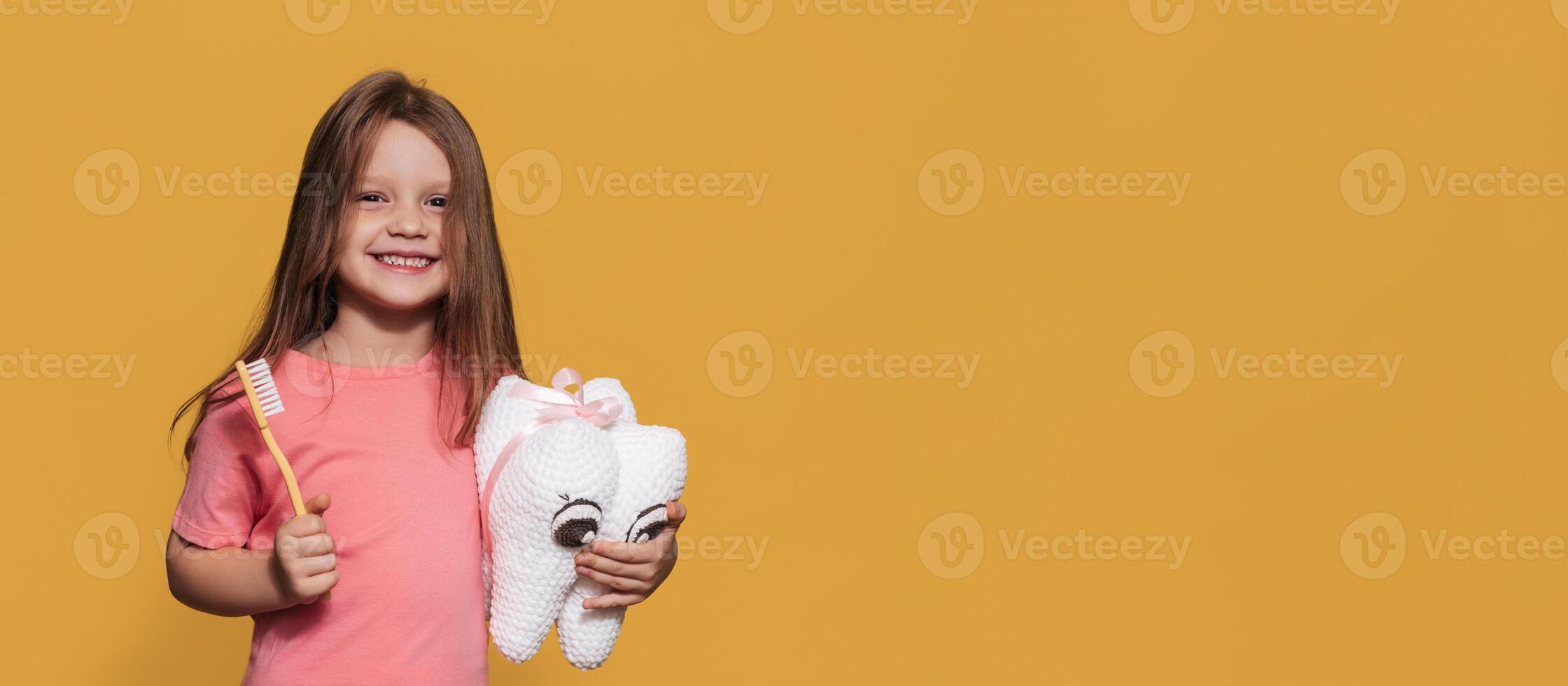 A smiling girl with healthy teeth holds a toy tooth and a toothbrush on a yellow isolated background. A place for your text. Oral hygiene. Pediatric dentistry. Prosthetics. Rules for brushing teeth. photo
