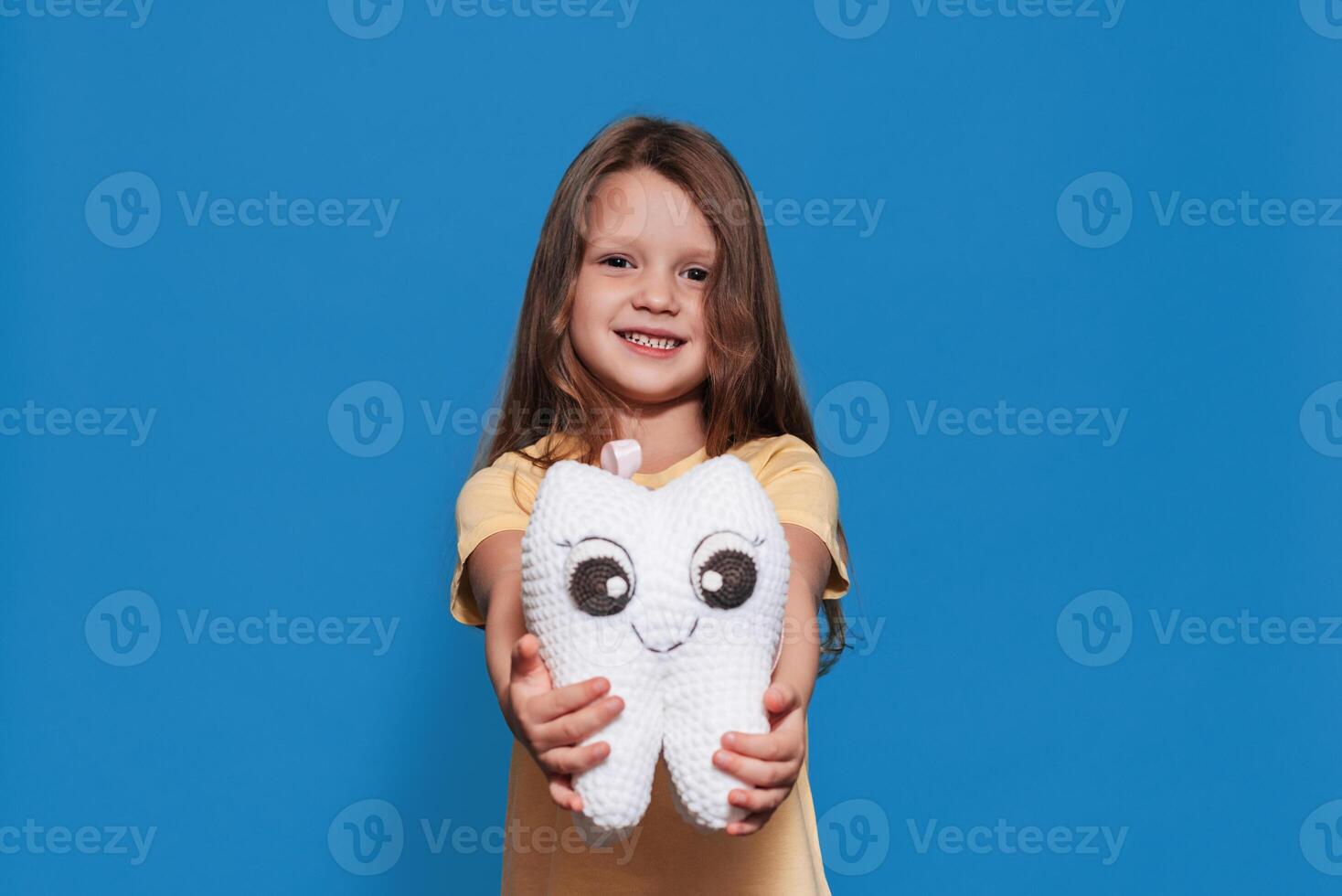 A smiling girl with healthy teeth holds a tooth toy in her hands on a blue isolated background. Oral hygiene. Pediatric dentistry. Prosthetics. Rules for brushing teeth. photo