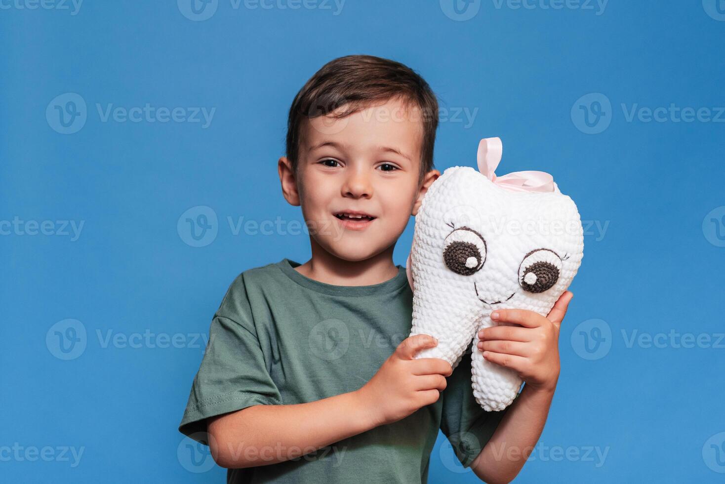 A smiling boy with healthy teeth holds a plush tooth on a blue background. Oral hygiene. Pediatric dentistry. photo
