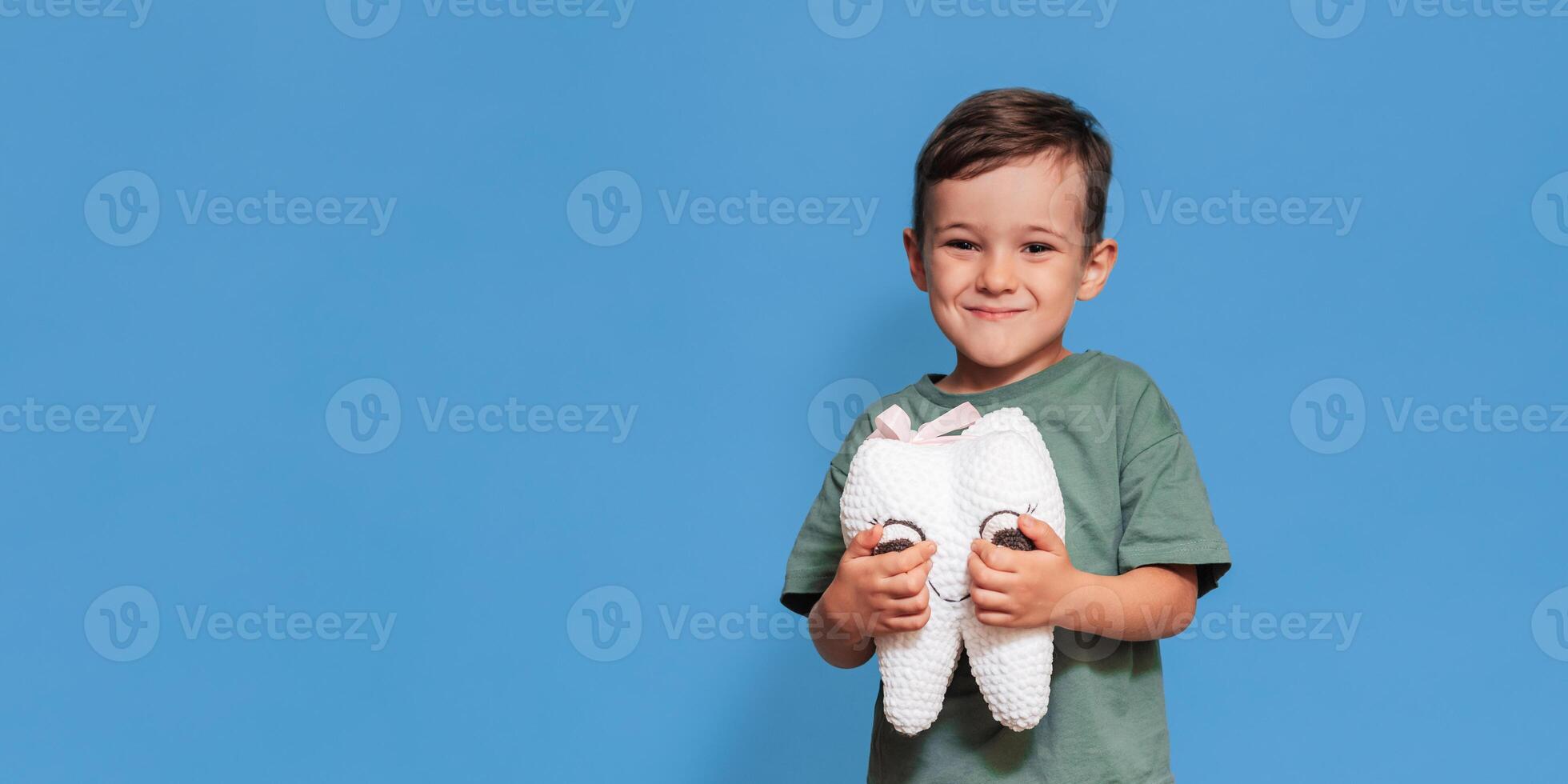 A smiling boy with healthy teeth holds a plush tooth in his hands on a blue isolated background. Oral hygiene. Pediatric dentistry. Prevention of caries. A place for your text. photo