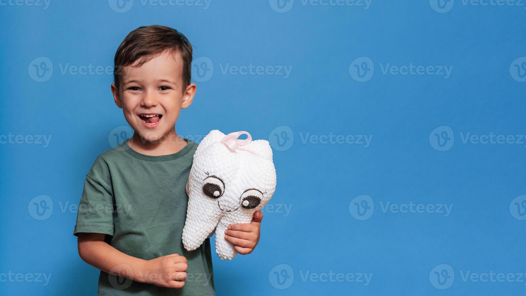 A smiling boy with healthy teeth holds a toothbrush and a plush tooth on a blue background. Pediatric dentistry. A place for your text. Panoramic photo. photo