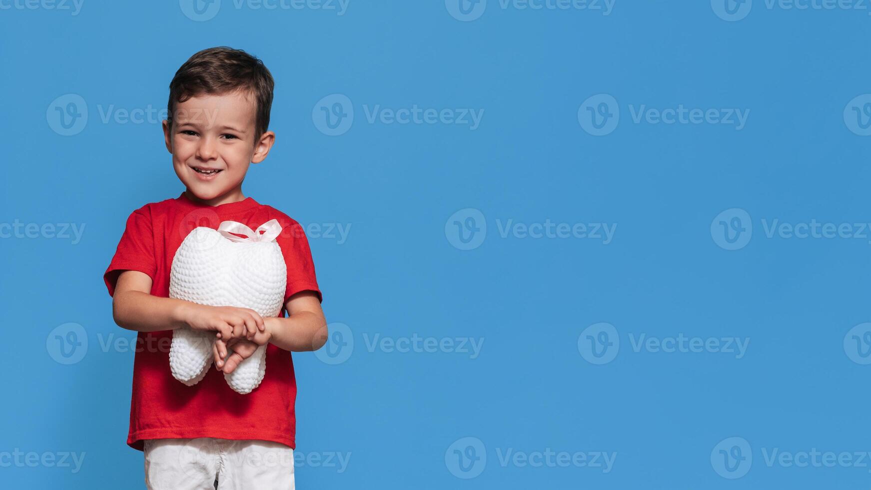 A smiling boy with healthy teeth holds a plush tooth in his hands on a blue isolated background. Oral hygiene. Pediatric dentistry. Prevention of caries. A place for your text. photo