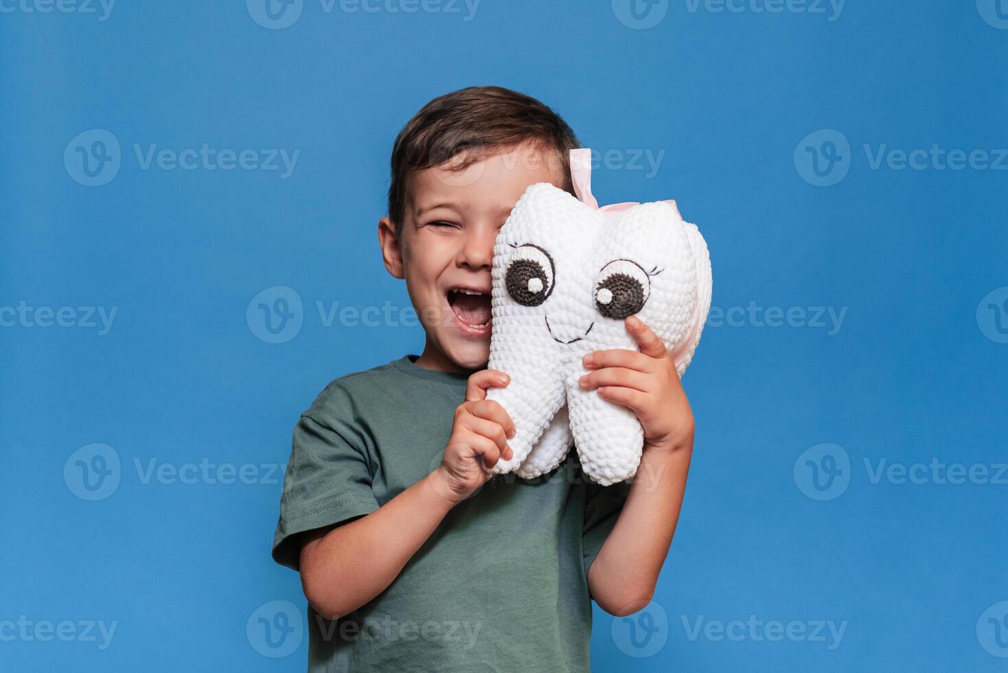 A smiling boy with healthy teeth holds a plush tooth on a blue background. Oral hygiene. Pediatric dentistry. photo