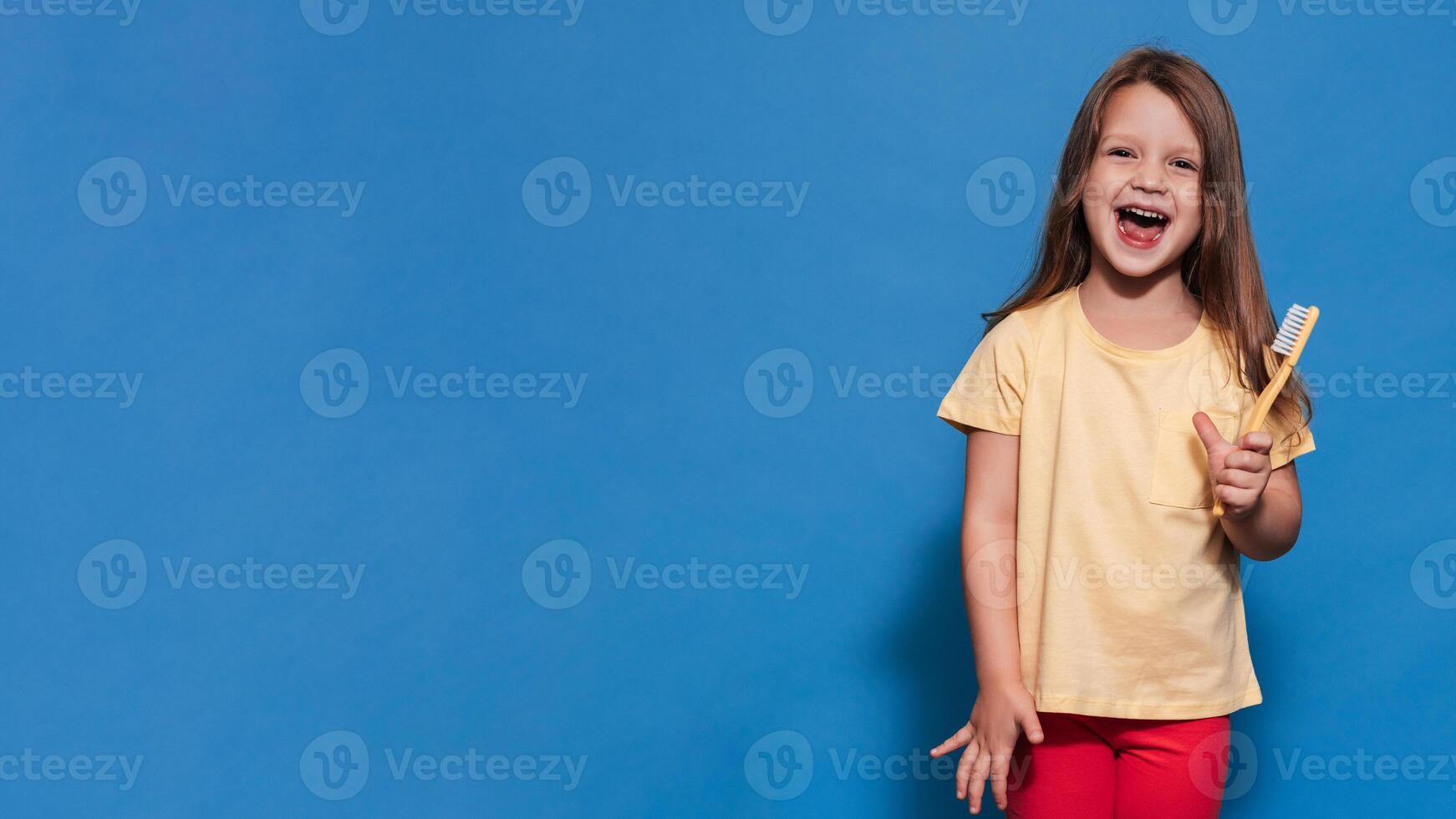 A smiling girl with healthy teeth holds a toothbrush in her hands on a yellow isolated background. A place for your text. Oral hygiene. Pediatric dentistry. Prosthetics. Rules for brushing teeth. photo