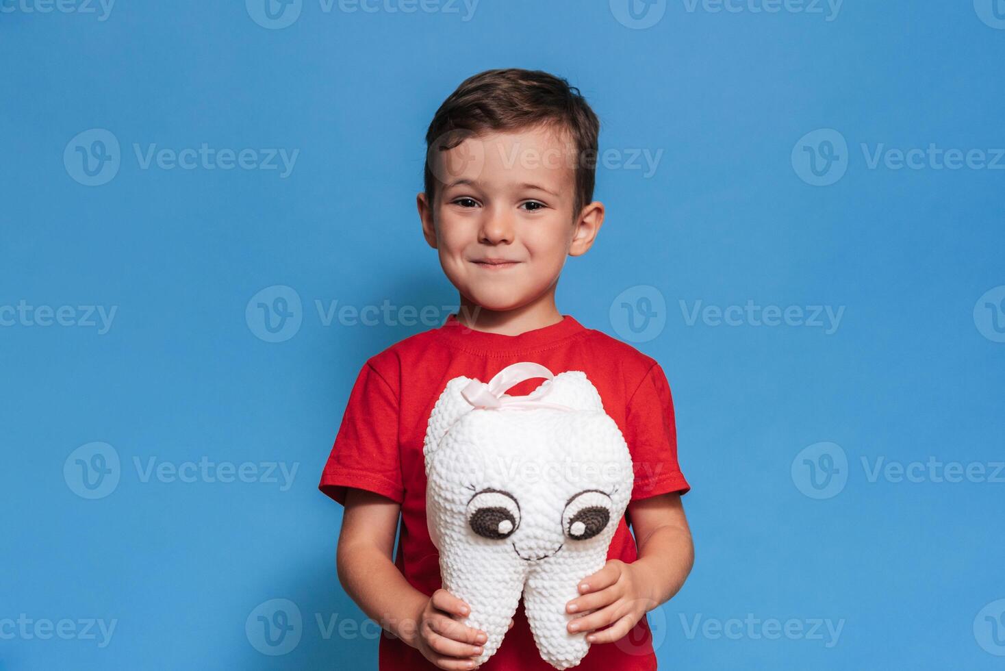 A smiling boy with healthy teeth holds a plush tooth in his hands on a blue isolated background. Oral hygiene. Pediatric dentistry. Prevention of caries. A place for your text. photo