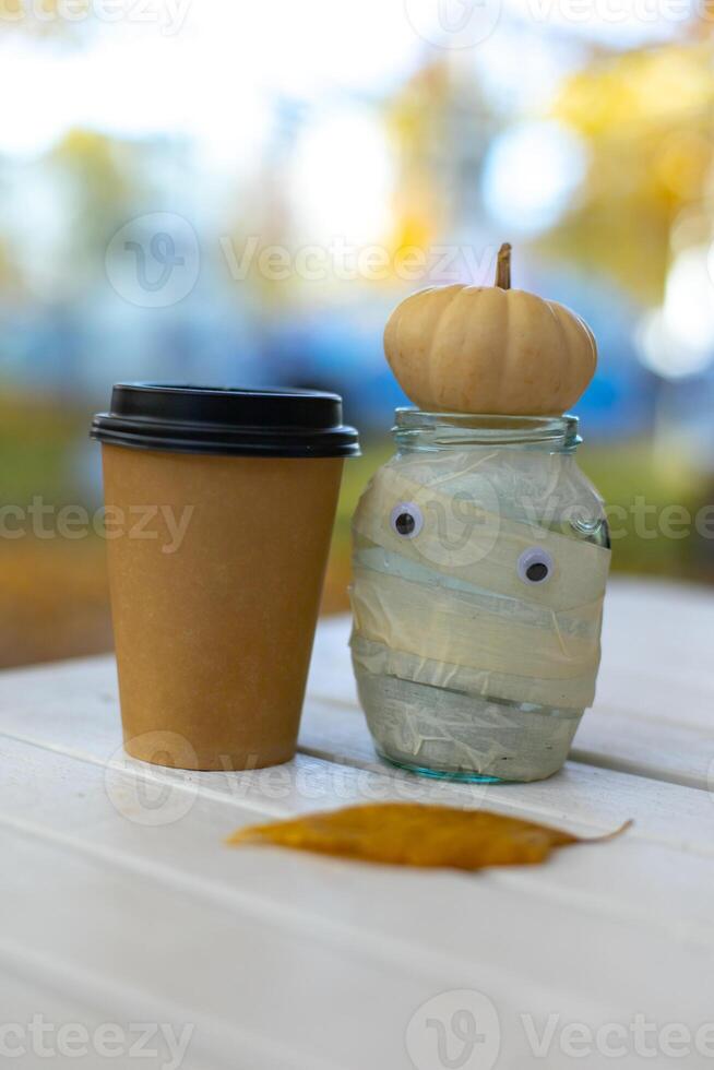 jar decor mummy with halloween pumpkin and craft glass with coffee on autumn bokeh background photo