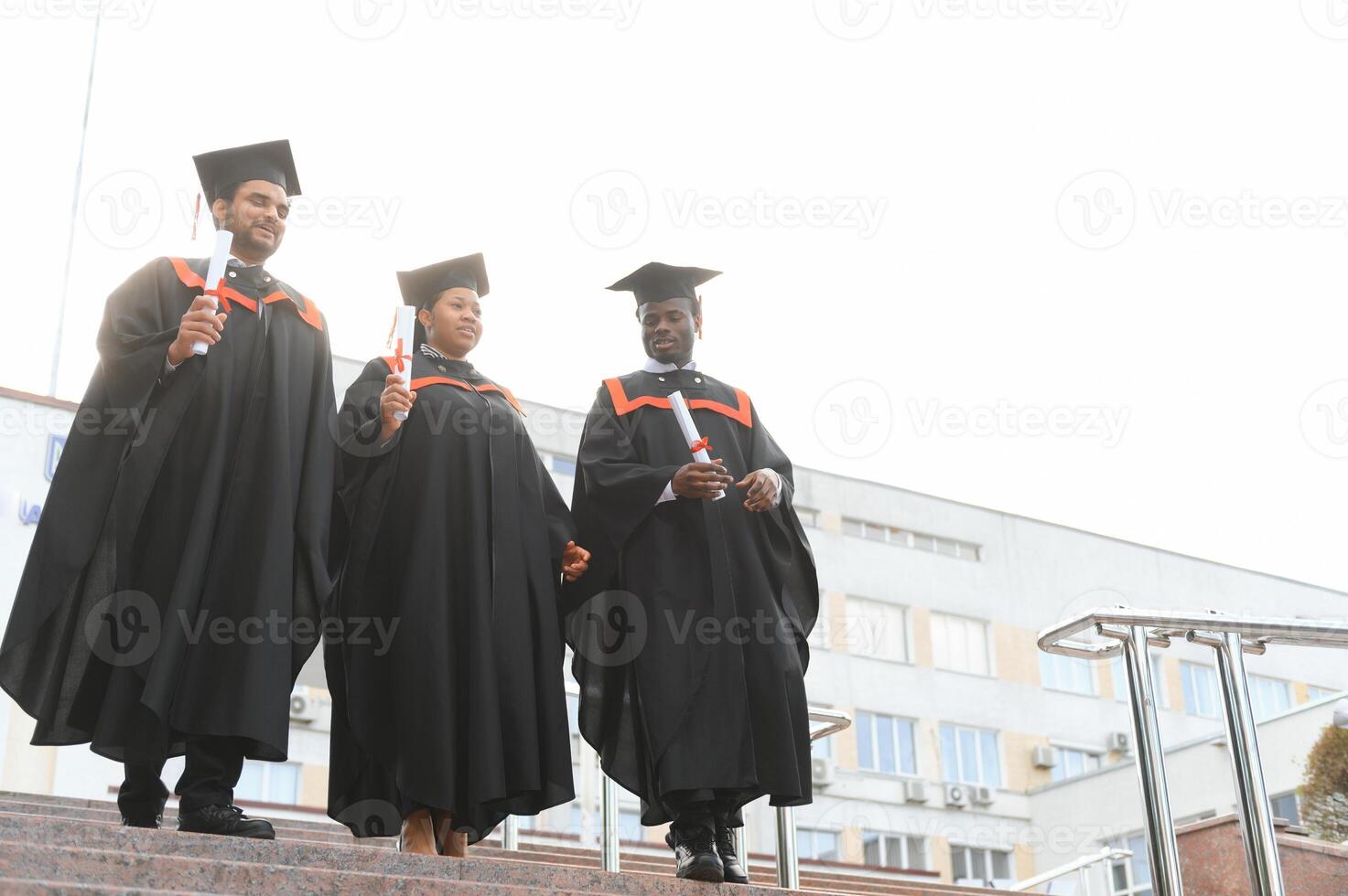 Group of happy smiling multicultural people in graduation gowns and caps with diplomas in hands graduate university outdoors in campus looking at camera. Graduation from college, university concept. photo