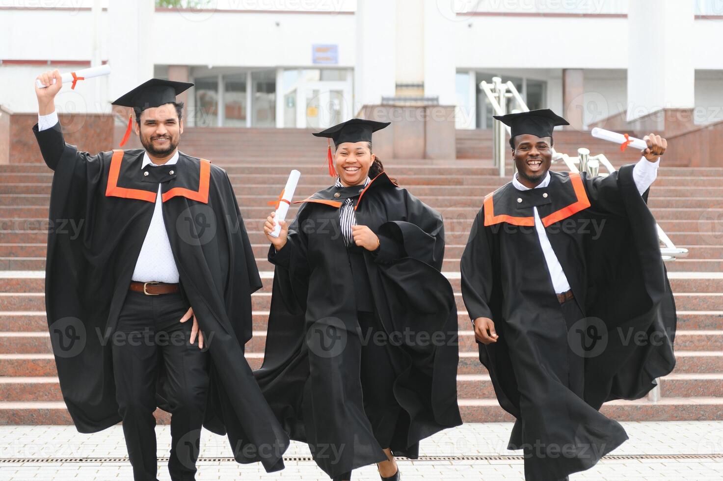 Group of happy smiling multicultural people in graduation gowns and caps with diplomas in hands graduate university outdoors in campus looking at camera. Graduation from college, university concept. photo