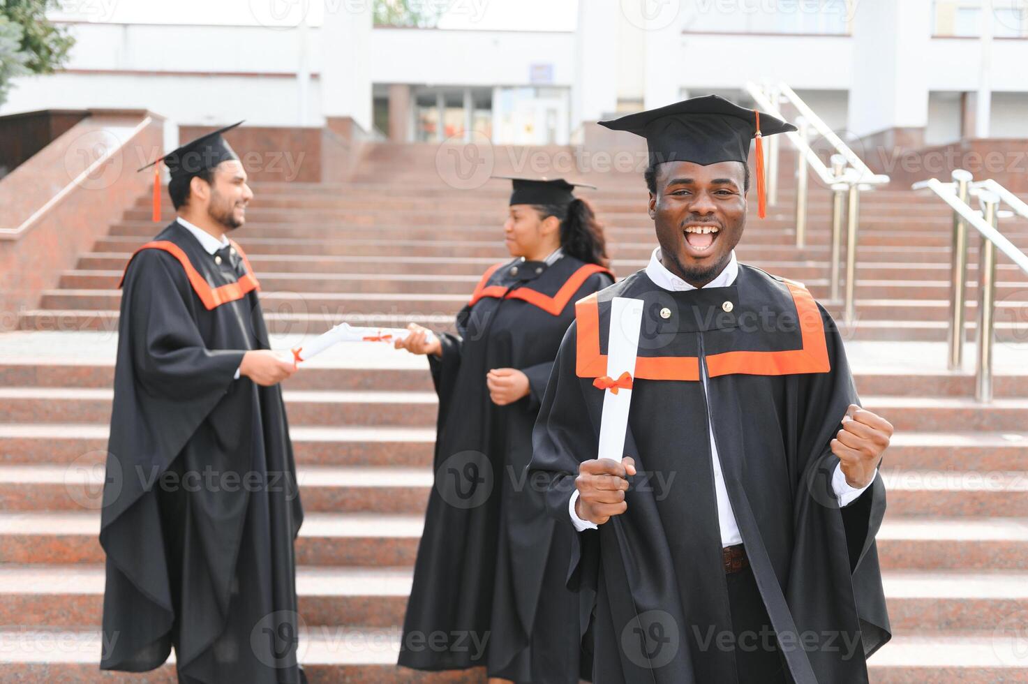 Happy graduate. Happy African man in graduation gowns holding diploma and smiling while his friends standing in the background photo