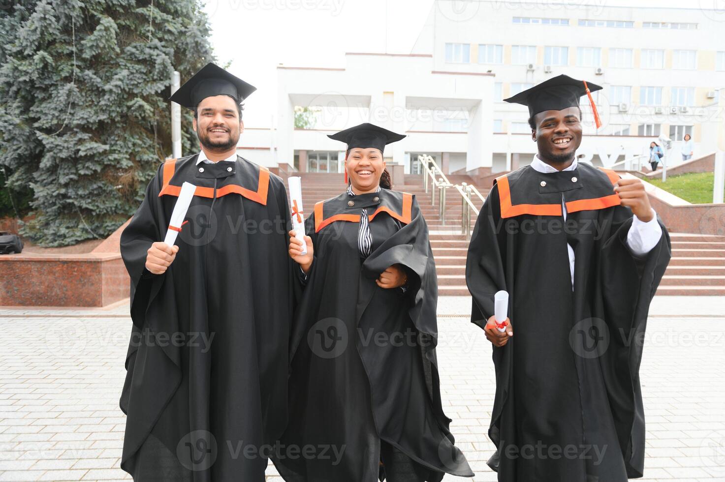 Group of happy smiling multicultural people in graduation gowns and caps with diplomas in hands graduate university outdoors in campus looking at camera. Graduation from college, university concept. photo