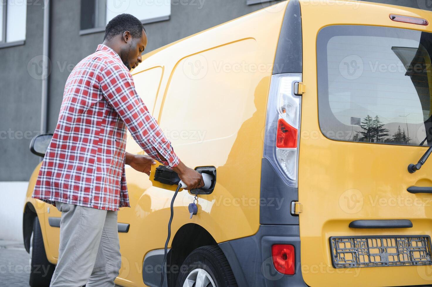 African american Man stands next to electric delivery vans at electric vehicle charging station photo