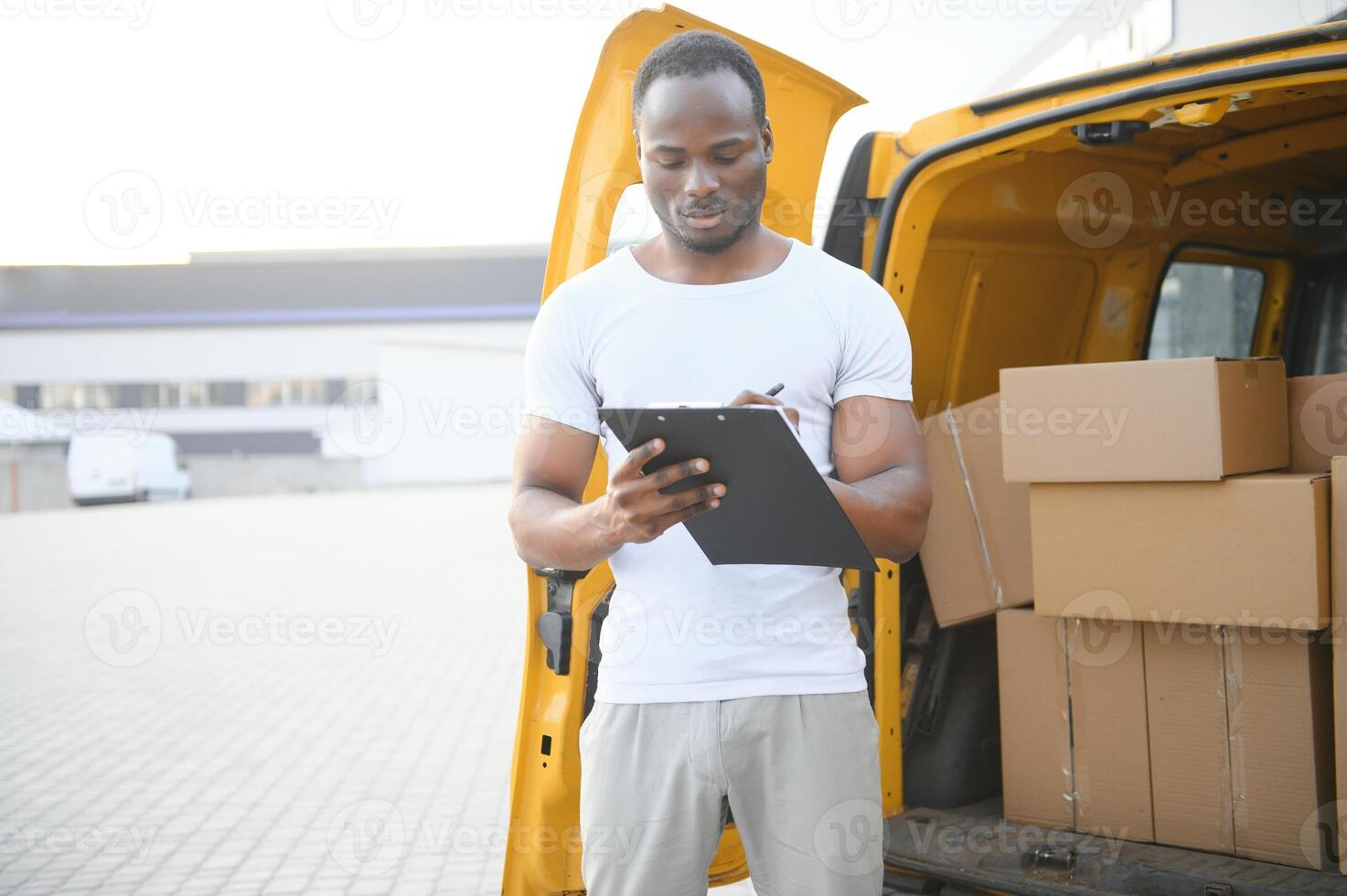 african american Worker unloading boxes from van outdoors. Moving service concept. Male employee of moving company makes inventory of packed boxes and writes data to clipboard. photo