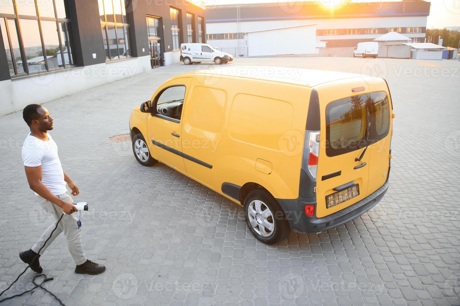 A african american man stands next to yellow electric delivery van at electric vehicle charging stations photo