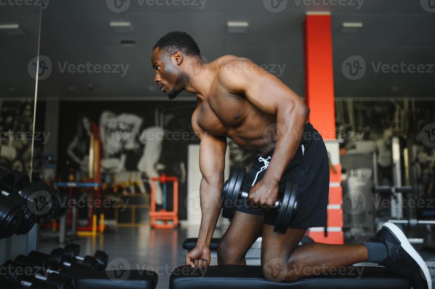 Young African American man sitting and lifting a dumbbell close to the rack at gym. Male weight training person doing a biceps curl in fitness center photo