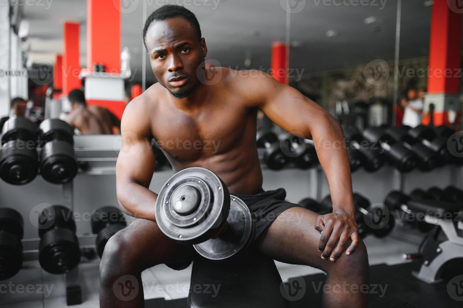 Young African American man sitting and lifting a dumbbell close to the rack at gym. Male weight training person doing a biceps curl in fitness center photo