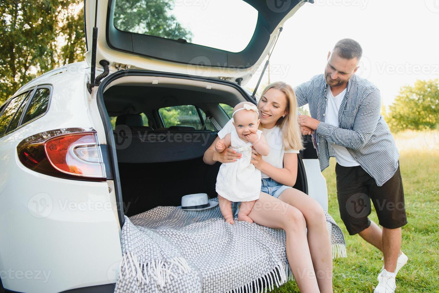 Pretty young married couple and their daughter are resting in the nature. The woman and girl are sitting on open car boot. The man is standing near them. They are smiling photo