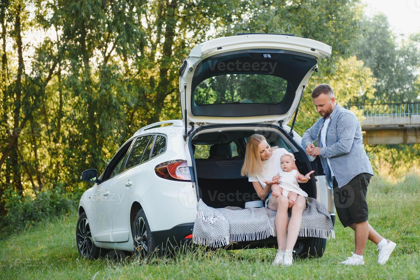 Pretty young married couple and their daughter are resting in the nature. The woman and girl are sitting on open car boot. The man is standing near them. They are smiling photo