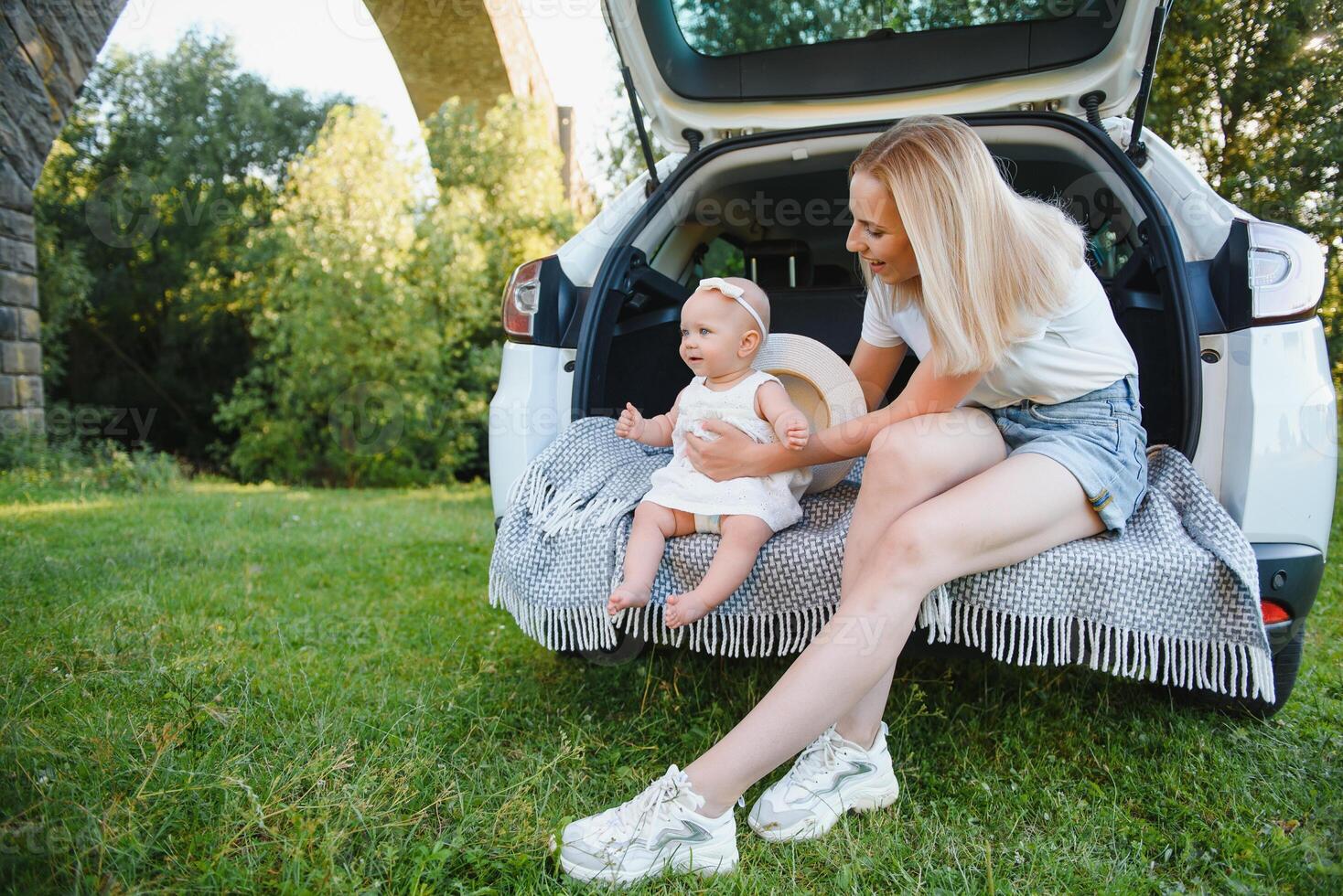 Pretty young married couple and their daughter are resting in the nature. The woman and girl are sitting on open car boot. photo