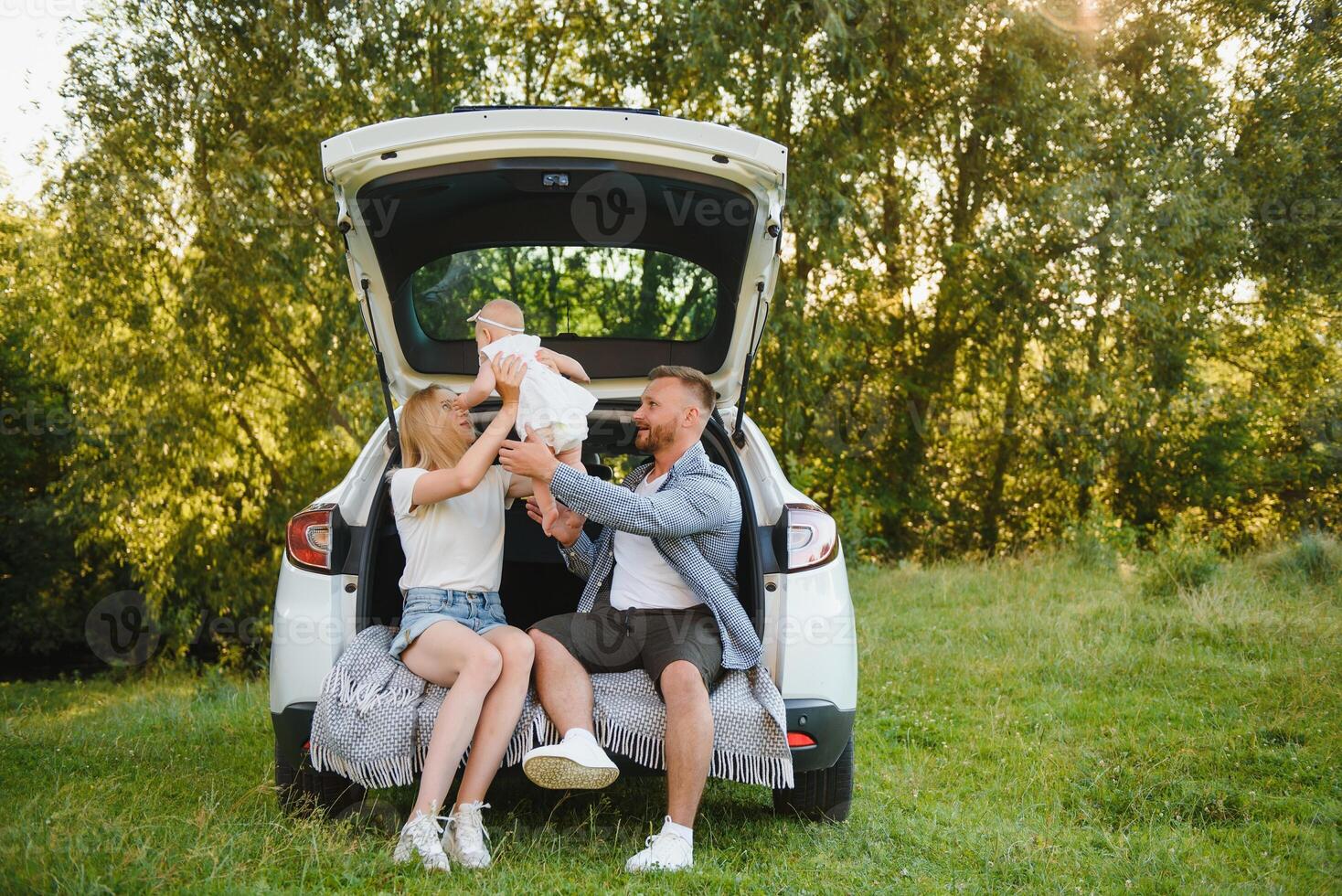 Pretty young married couple and their daughter are resting in the nature. The woman and girl are sitting on open car boot. photo