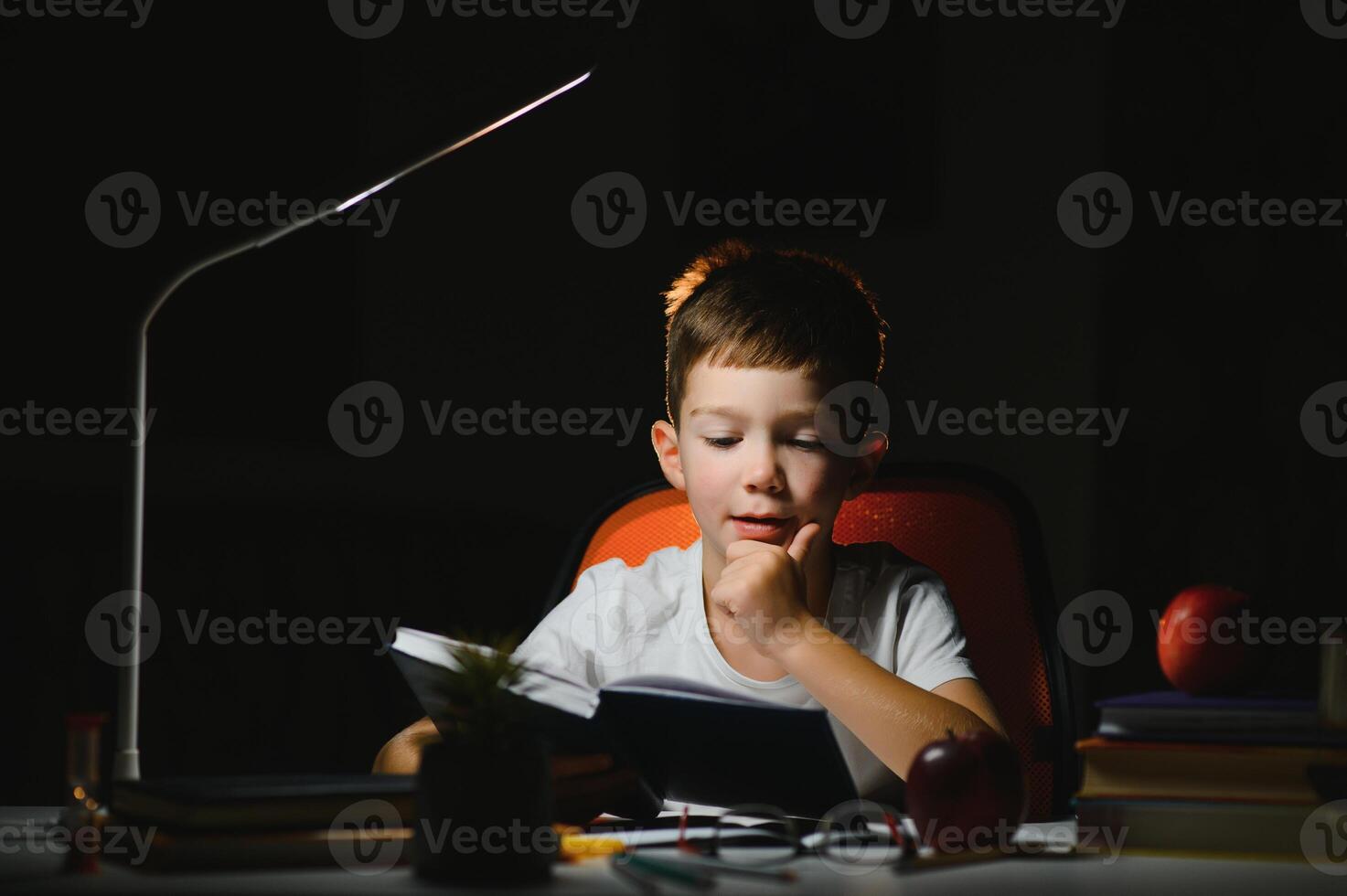 concentrated schoolboy reading book at table with books, plant, lamp, colour pencils, apple, and textbook photo