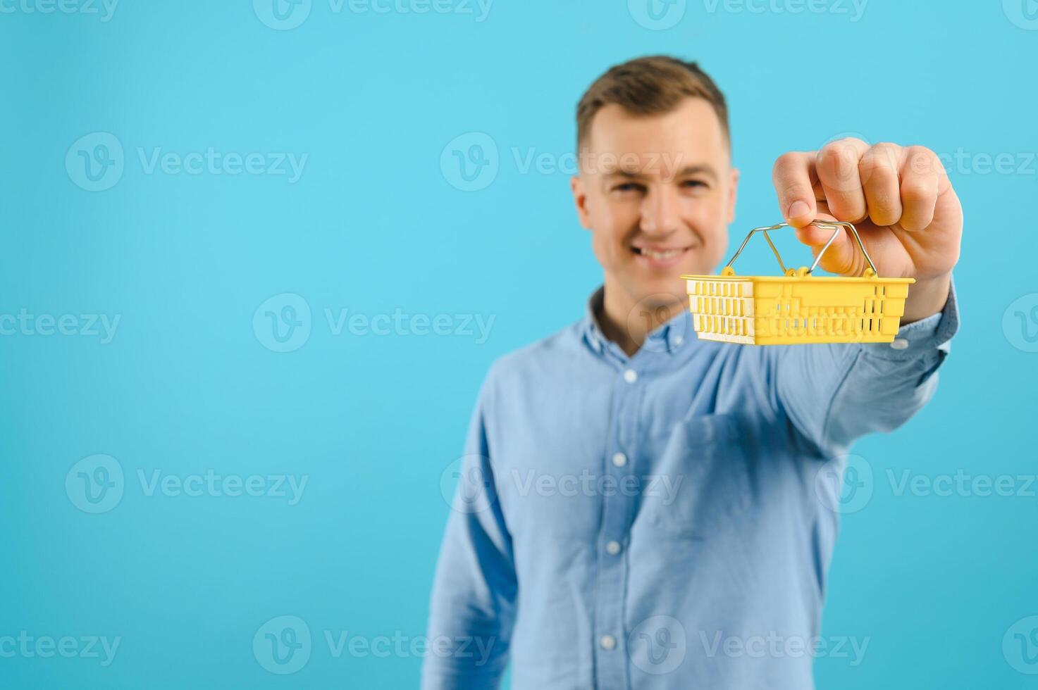 A young man holding a mini shopping cart and standing in front of a blue background in the studio. Sales concept. Place for text. photo